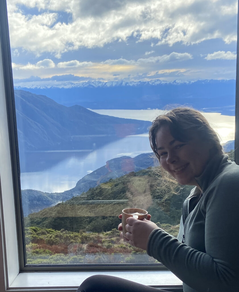 Ella Dewar enjoying a hot drink by a window with a few over lake Te Anau.